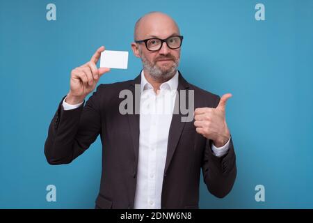 Caucasien mature homme dans le costume d'affaires montrant les pouces vers le haut signe montrant la carte de crédit. studio tourné sur le mur bleu Banque D'Images