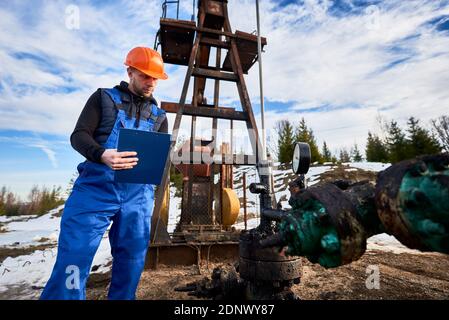 Ingénieur sérieux dans les combinaisons de travail et le casque tenant le presse-papiers et regardant la machine à bascule de pompe à huile. Employé vérifiant l'unité de pompage d'huile, prenant des notes. Concept de l'extraction de pétrole et de l'industrie pétrolière Banque D'Images