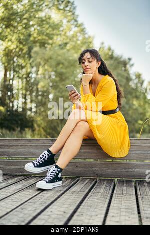 Jeune femme portant robe jaune et noir sneakers et à l'aide du smartphone sur le trottoir de bois Banque D'Images