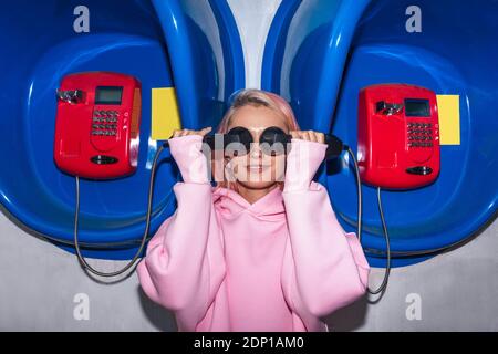 Jeune femme souriante aux cheveux roses portant une chemise à capuchon rose debout dans les cabines téléphoniques, tenant des récepteurs devant les yeux Banque D'Images