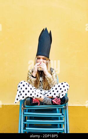 Portrait of Girl wearing black crown assis sur pile de chaises eating Hamburger Banque D'Images