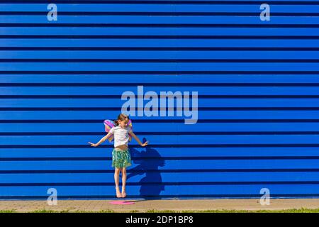 Petite fille avec des ailes de papillon colorées sautant dans l'air devant un arrière-plan bleu Banque D'Images