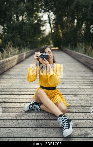 Jeune femme photographe portant robe jaune et noir sneakers tenant une caméra analogique sur trottoir de bois Banque D'Images