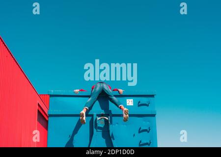 Jeune homme suspendu sur le bord du conteneur bleu, vue arrière Banque D'Images