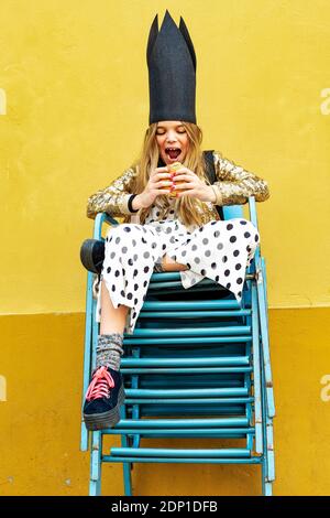 Portrait of Girl wearing black crown assis sur pile de chaises eating Hamburger Banque D'Images
