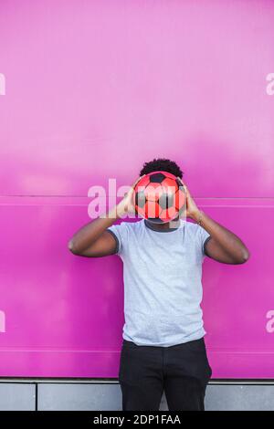 Jeune homme tenant le ballon de football devant le visage pendant se tenant contre la couleur rose Banque D'Images