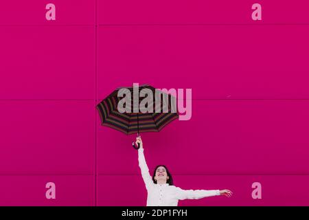 Jeune femme sautant avec un parapluie devant un rose mur Banque D'Images