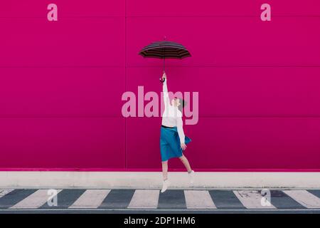 Jeune femme sautant avec un parapluie devant un rose mur Banque D'Images
