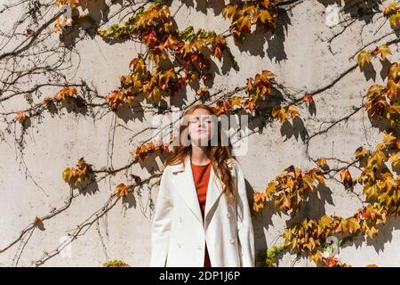 Jeune femme à tête rouge portant une veste debout avec les yeux fermés contre mur ivy Banque D'Images