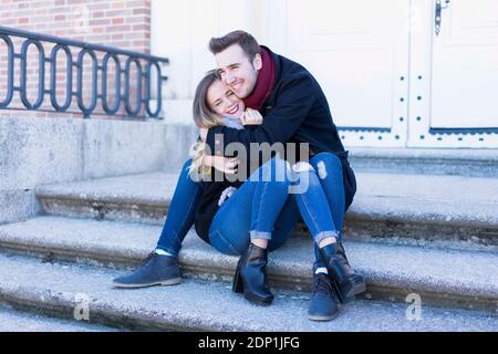 Portrait d'un couple souriant dans des vêtements d'hiver qui s'embrasent assis quelques escaliers sur une scène d'hiver de parc urbain Banque D'Images