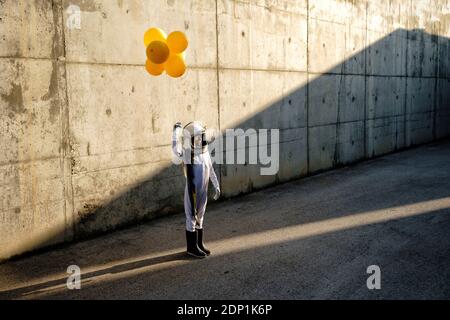 Petite fille portant un casque d'espace tenant un ballon debout dans la rue contre le mur Banque D'Images