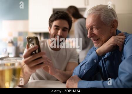 Père âgé souriant et regardant des photos sur smartphone de Happy jeune homme assis dans la cuisine pendant le dîner en famille Banque D'Images