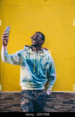 Portrait of smiling young man taking smartphone selfies avec en face de mur jaune Banque D'Images