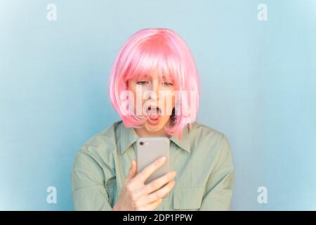 Portrait d'une jeune femme surprise portant une perruque rose regardant téléphone portable Banque D'Images