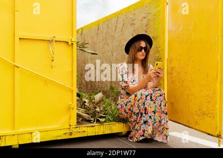 Femme portant une robe fleurie, assise dans un récipient jaune, utilisant un smartphone Banque D'Images
