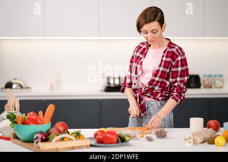 Couper des carottes fraîches maison cuisine ragoût de viande ou ragoût portant une chemise à carreaux. Cuisiner avec passion jeune femme aux cheveux courts debout à la moderne Banque D'Images