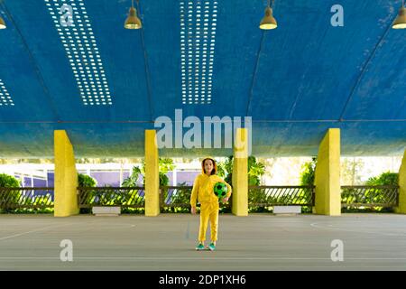 Petite fille portant un survêtement jaune tenant un ballon de football sous elle bras Banque D'Images