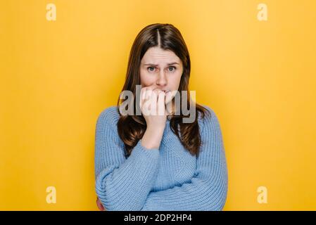 Gros plan portrait de la jeune femme du caucase nerveux piquant ses ongles tout en regardant la caméra, vêtue de chandail bleu tricoté, isolé sur stu jaune Banque D'Images