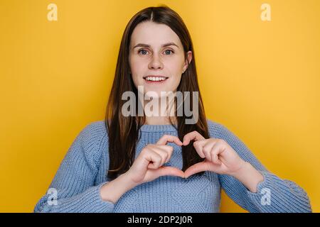Gai mignon jeune femme ayant le large sourire de charme mettre les mains sur la poitrine faire avec les doigts coeur forme symbole de la charité, isolé sur le studio jaune Banque D'Images