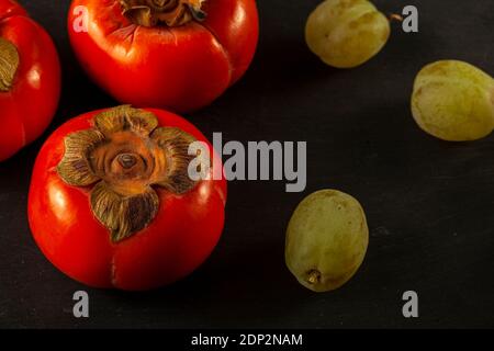 Gros plan de quelques fruits Sharon (persimmons) et quelques raisins verts sur fond noir en bois. Une composition simple de saine alimentation fraîche co Banque D'Images