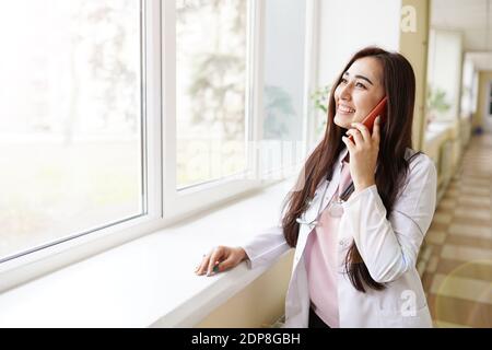 Bonne belle jeune femme médecin debout et parlant sur la cellule téléphone près de la fenêtre à l'hôpital Banque D'Images