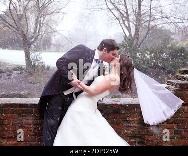 Neige tombant lors d'un mariage enneigé avec mariée et marié embrassant. La mariée porte une robe de mariage blanche avec voile. Le marié porte un costume aux queues. Banque D'Images
