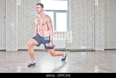 Jeune homme beau avec une construction sportive faisant des fentes avec des haltères dans le hall avec une fenêtre. Style de vie sportif Banque D'Images