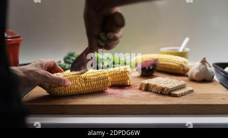 Vue à angle bas des mains femelles coupant des épis de maïs sur une planche à découper en bois pleine de légumes et de protéines végétaliennes, tempeh. Banque D'Images