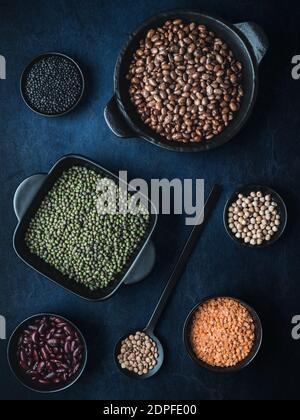 Bols noirs de différentes légumineuses séchées - haricots, lentilles, haricots mungo et pois chiches sur fond bleu foncé. Vue aérienne. Sombre et moody. Banque D'Images
