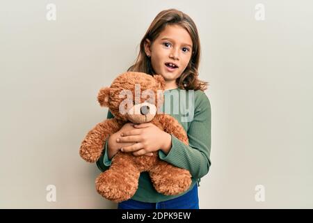 Petite belle fille embrassant l'ours en peluche dans le visage de choc, regardant sceptique et sarcastique, surpris par la bouche ouverte Banque D'Images