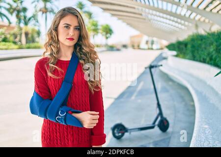 Jeune fille blonde d'expression sérieuse injuriée avec une élingue de bras debout au parc. Banque D'Images