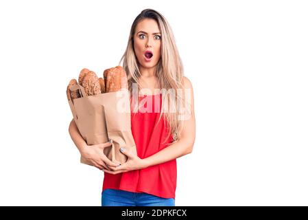 Jeune belle femme blonde tenant le sac de papier avec du pain effrayé et stupéfié avec la bouche ouverte pour la surprise, le visage incrédulé Banque D'Images