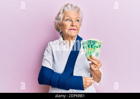 Femme sénior à cheveux gris portant le bras sur une élingue tenant des billets de banque israéliens souriant regardant sur le côté et regardant loin de penser. Banque D'Images