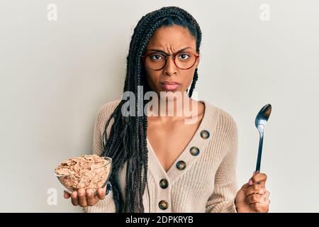 Femme afro-américaine avec des tresses mangeant des céréales complètes saines sceptique et nerveux, frowning bouleversé à cause du problème. Personne négative. Banque D'Images