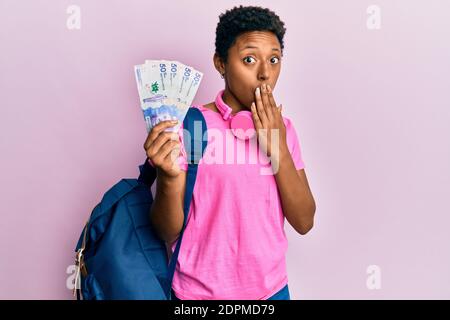 Une jeune fille afro-américaine portant un sac à dos étudiant portant des billets colombiens en pesos couvrant la bouche avec la main, choquée et effrayée par erreur Banque D'Images