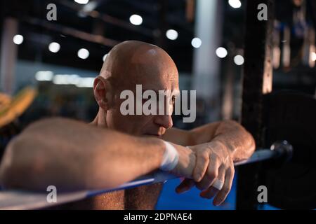 Athlète âgé bald se pentant sur la barbell et se reposant pendant la pause dans l'exercice fonctionnel intense dans la salle de gym Banque D'Images