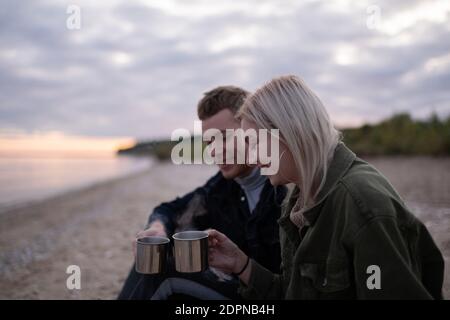 Vue latérale d'un jeune homme et d'une femme souriant et heureux prendre des tasses de thé chaud tout en se reposant sur la rive de l'étang en soirée nuageux Banque D'Images