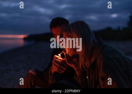 Jeune homme éclairant la cigarette de cannabis pour petite amie pendant qu'il est assis automne nature et repos la nuit Banque D'Images