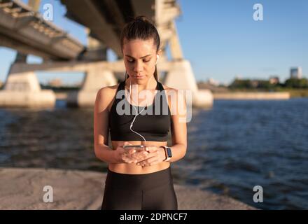 Jeune femme concentrée dans une tenue sportive noire portant des écouteurs et utilisation du smartphone pour préparer la liste de lecture de musique pour l'entraînement en ville front de mer Banque D'Images