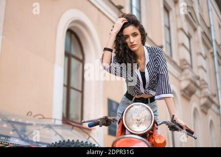 Jeune femme brune en chemise rayée et jeans assis sur une moto rouge à l'extérieur. Banque D'Images