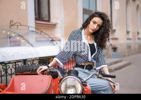 Jeune femme brune en chemise rayée et jeans assis sur une moto rouge à l'extérieur. Banque D'Images