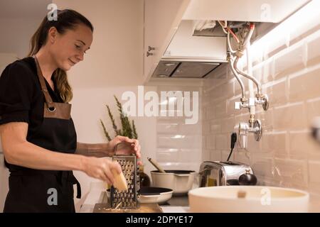 Jeune belle femme debout près du poêle et râper le fromage pour une recette saine de nourriture. Charmante femme norvégienne travaillant comme blogueur de nourriture et lif Banque D'Images