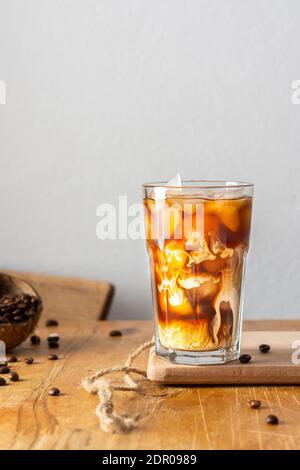 Café froid rafraîchissant avec glace et nuages crémeux dans un verre sur une planche en bois gros plan, fèves de cacao dans la noix de coco Banque D'Images