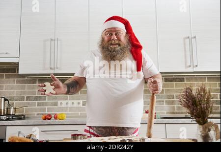 Un homme barbu joyeux dans les tatouages et un père noël chapeau tient d'une main la forme d'un pain d'épice homme fait de pâte et dans l'autre un bois roulement Banque D'Images