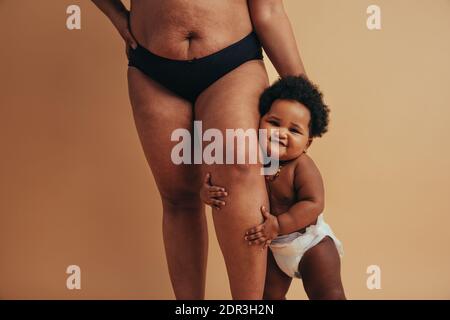 Bébé mignon accroché à la jambe de sa mère. Plan court d'une femme avec le corps post-partum debout avec son bébé. Banque D'Images