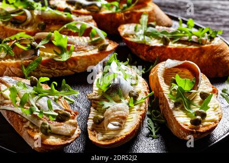 gros plan de sandwiches ouverts au beurre d'anchois avec feuilles d'arugula fraîches, câpres sur un plateau noir sur une table en bois rustique, vue horizontale depuis le dessus Banque D'Images