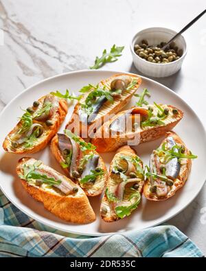 Bruschetta au beurre d'anchois, feuilles de salade de roquette fraîches, câpres sur un plateau sur une table en marbre, vue horizontale depuis le dessus Banque D'Images