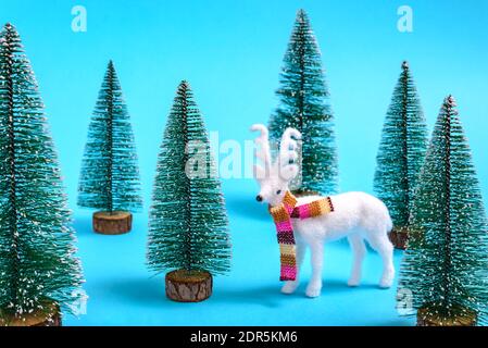 Concept créatif hiver et Noël de renne blanc avec foulard coloré dans une forêt de sapins sur fond bleu pastel. Banque D'Images