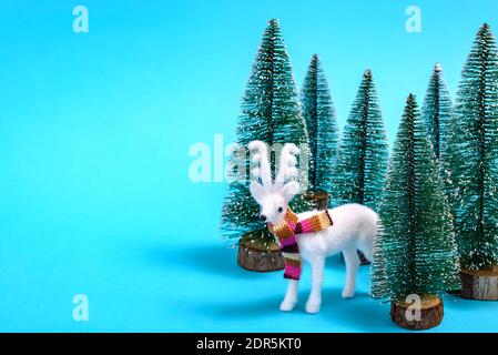 Concept créatif hiver et Noël de renne blanc avec foulard coloré dans une forêt de sapins sur fond bleu pastel. Banque D'Images