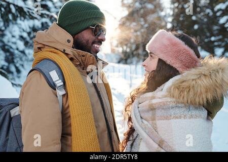 Vue latérale des jeunes heureux dates interculturelles en hiver l'un à l'autre devant l'appareil photo pendant le refroidissement forêt le week-end d'hiver Banque D'Images